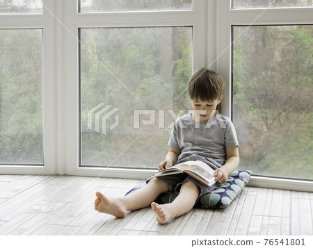 Kid sits on floor and reads book. Intellectual leisure for children while it's raining outside. 76541801