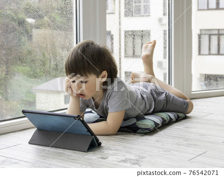 Curious boy watch cartoons on digital tablet. Kid lies on floor and uses electronic device. Indoor leisure for children while it's raining outside. 76542071