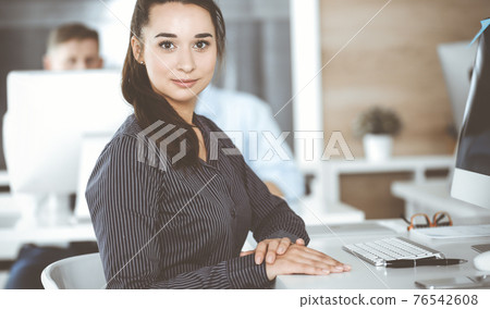 Business woman using computer at workplace in modern office. Brunette secretary or female lawyer looking at the camera and happy smiling. Working for pleasure and success Business woman using computer at workplace in modern office. Brunette secretary or female lawyer looking at the camera and happy smiling. Working for pleasure and success 76542608
