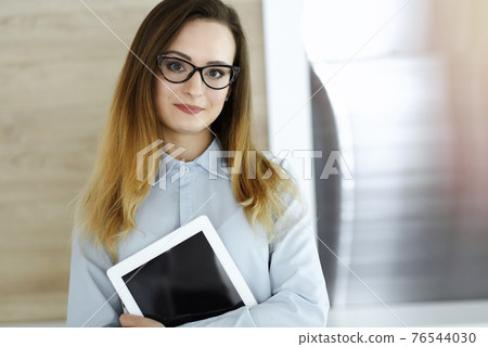 Business woman headshot in sunny office. Unknown businesswoman standing straight with tablet computer. Young accountant or secretary looks good Business woman headshot in sunny office. Unknown businesswoman standing straight with tablet computer. Young accountant or secretary looks good 76544030