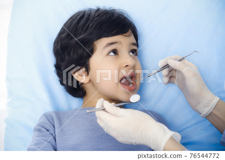Cute arab boy sitting at dental chair with open mouth during oral checking up with doctor. Visiting dentist office. Stomatology concept Cute arab boy sitting at dental chair with open mouth during oral checking up with doctor. Visiting dentist office. Stomatology concept 76544772