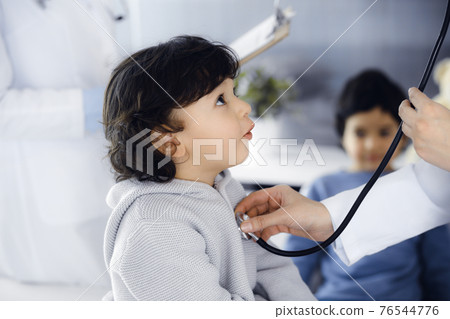 Woman-doctor examining a child patient by stethoscope. Cute arab toddler and his brother at physician appointment. Medicine concept Woman-doctor examining a child patient by stethoscope. Cute arab toddler and his brother at physician appointment. Medicine concept 76544776