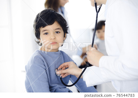 Doctor-woman examining a child patient by stethoscope. Cute arab boy and his brother at physician appointment. Medicine concept Doctor-woman examining a child patient by stethoscope. Cute arab boy and his brother at physician appointment. Medicine concept 76544783