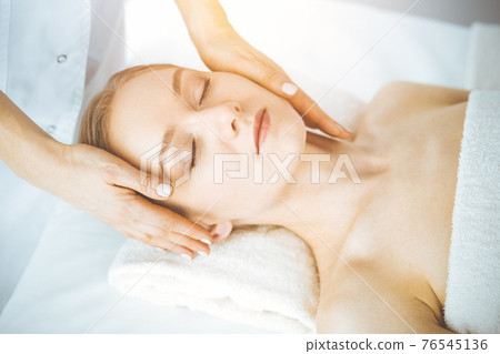 Happy woman enjoying facial massage with closed eyes in spa salon. Relaxing treatment in medicine and Beauty concept 76545136
