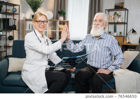 Cheerful positive blond woman doctor satisfied from successful treatment and result of x-ray CT scan of her retired male patient and giving high five each other during home visit Cheerful positive blond woman doctor satisfied from successful treatment and result of x-ray CT scan of her retired male patient and giving high five each other during home visit 76546650