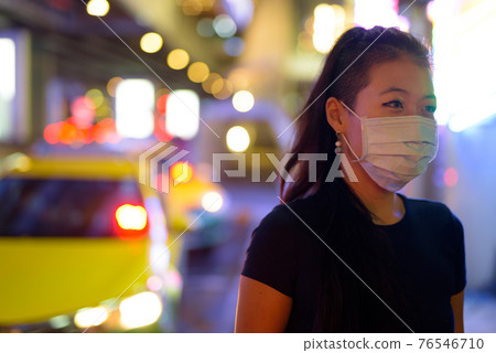 Asian woman wearing face mask to protect from covid 19 outdoors at night while at taxi station Asian woman wearing face mask to protect from covid 19 outdoors at night while at taxi station 76546710