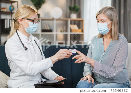 Blond female doctor in medical coat, glasses and protective mask, measuring oxygen saturation and heart rate with a pulse oximeter, while visiting her sick retired lady patient at home 76546882