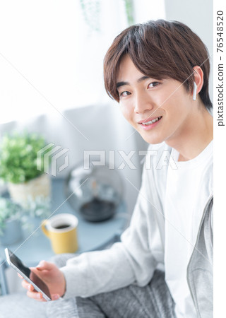 A young man holding a smartphone in his living room 76548520
