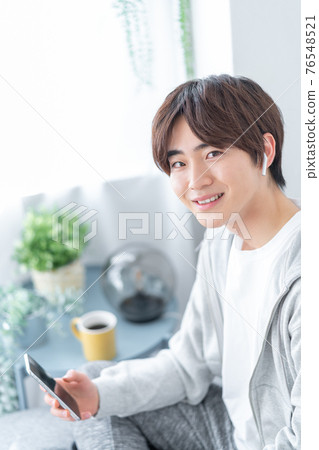 A young man holding a smartphone in his living room 76548521