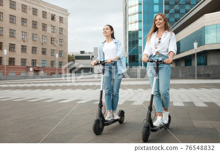 Female friends riding motorized kick scooters in the city Female friends riding motorized kick scooters in the city 76548832