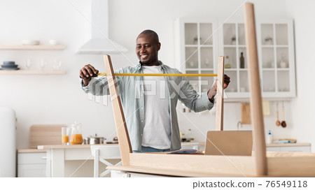 Excited black man measuring furniture with roulette, assembling new table in kitchen interior Excited black man measuring furniture with roulette, assembling new table in kitchen interior 76549118