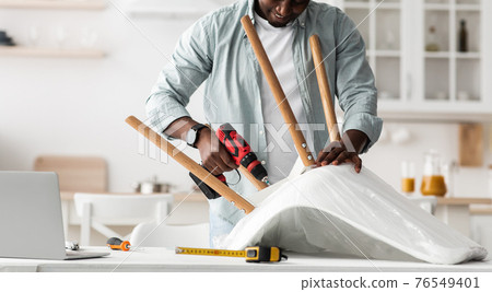 Repair or furniture assembly process. Craftsman screwing legs to chair with electric screwdriver in kitchen, panorama Repair or furniture assembly process. Craftsman screwing legs to chair with electric screwdriver in kitchen, panorama 76549401