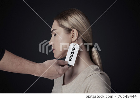 Physiology, mental problems, sexism and humiliation. Male hand hangs label with inscription frigid on upset young lady Physiology, mental problems, sexism and humiliation. Male hand hangs label with inscription frigid on upset young lady 76549444