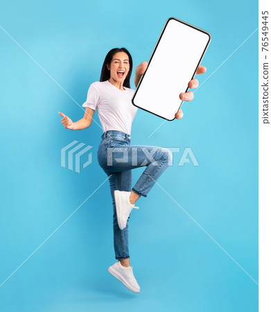 Advertisement for mobile app. Excited lady jumping over blue background, showing modern smartphone with empty screen 76549445