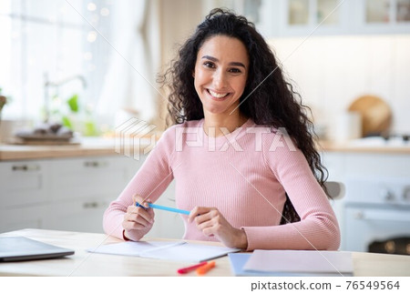 Smiling young woman sitting at table with workbooks and laptop in kitchen 76549564