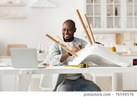 Happy african american man twisting part in chair and looking at laptop, watching online video instructions in kitchen Happy african american man twisting part in chair and looking at laptop, watching online video instructions in kitchen 76549576