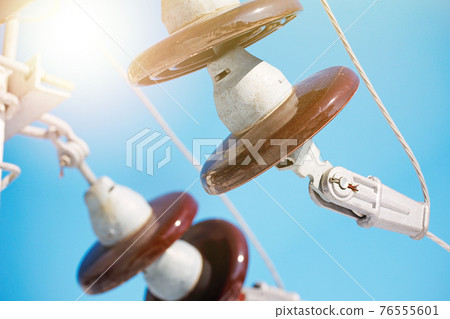 A ceramic suspended insulator hangs from a power line against a blue sky. A ceramic suspended insulator hangs from a power line against a blue sky. 76555601