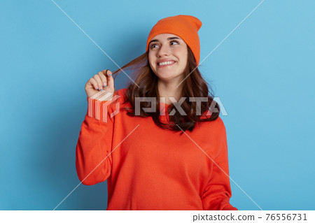 Young beautiful lady dreamily looks aside, smiling happily isolated over blue background, pulling her hair to side, wearing orange casual jumper and cap, having satisfied facial expression. 76556731