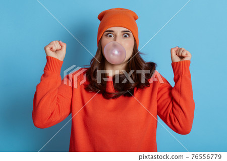 Excited dark haired female blows bubble from chewing gum, has amazed expression, dresses casual clothes, model posing isolated over blue background, cant believe in her success, clenches fists. 76556779