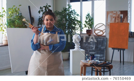 A woman painter posing with brushes in hand and looking in the camera A woman painter posing with brushes in hand and looking in the camera 76558469