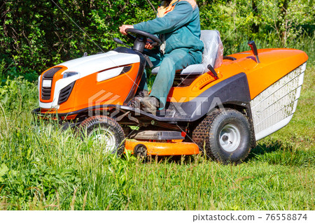A gardener mows a green meadow with a tractor lawn mower. 76558874