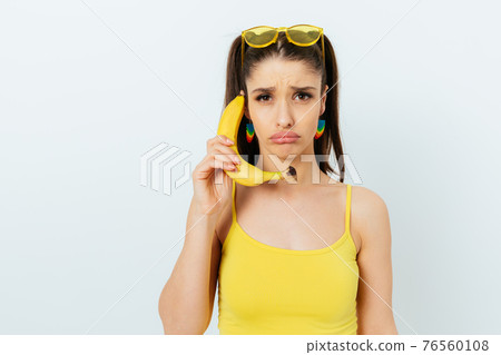Sad young woman wearing summer outfit holding banana 76560108