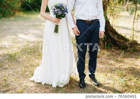 Bride with a bouquet of blue flowers and the groom stand side by side in the olive grove and hold hands 76560491