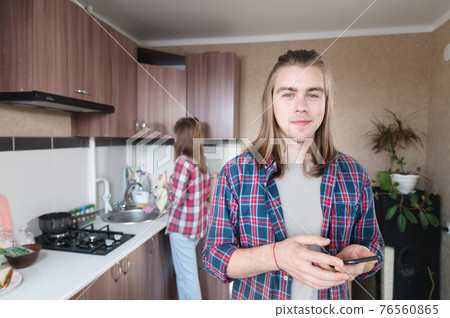 in the kitchen room, a young man with long hair stands with a phone in his hands and looks at the camera in the kitchen room, a young man with long hair stands with a phone in his hands and looks at the camera 76560865