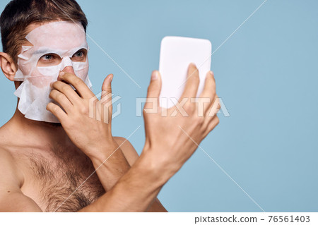 a man with a cleansing mask on his face examines himself in a mirror on a blue background 76561403