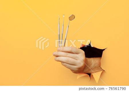 Hand showing a dentist tools out of a hole torn in yellow wall. Health care concept 76561790