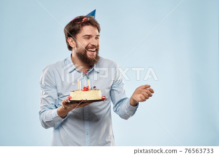Bearded man with a plate of cake on a blue background and a birthday party hat on his head Bearded man with a plate of cake on a blue background and a birthday party hat on his head 76563733