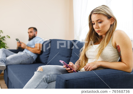 Scolded couple sitting on opposite sides of the couch using their smartphones, don't care for each other Scolded couple sitting on opposite sides of the couch using their smartphones, don't care for each other 76568736