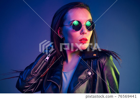 Asian girl in sunglasses and black leather jacket posing against dark blue background Asian girl in sunglasses and black leather jacket posing against dark blue background 76569441