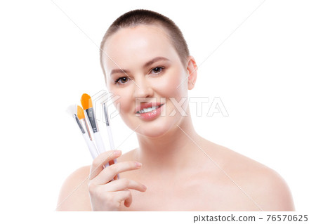Short-haired woman with bare shoulders showing make-up brushes set against white background 76570625