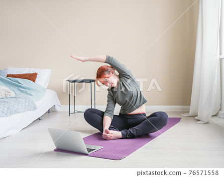 Red hair woman sitting on yoga mat near bed. Girl Red hair woman sitting on yoga mat near bed. Girl 76571558