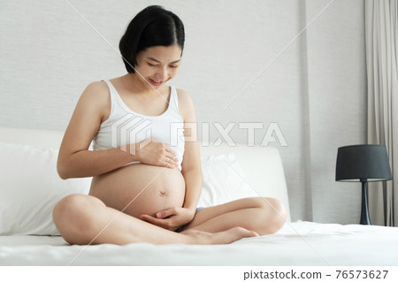 Cheerful Asian Young pregnant woman smiling sitting touching her beautiful belly on bed at home. Cheerful Asian Young pregnant woman smiling sitting touching her beautiful belly on bed at home. 76573627