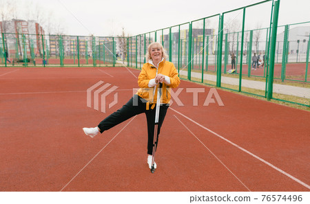 an elderly woman holding a Nordic walking stick stands on the red treadmill in the stadium 76574496