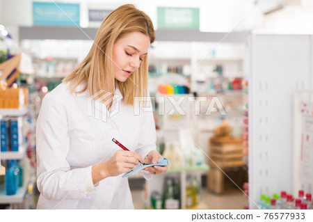 Diligent friendly smiling female pharmacist noting assortment of drugs in pharmacy Diligent friendly smiling female pharmacist noting assortment of drugs in pharmacy 76577933