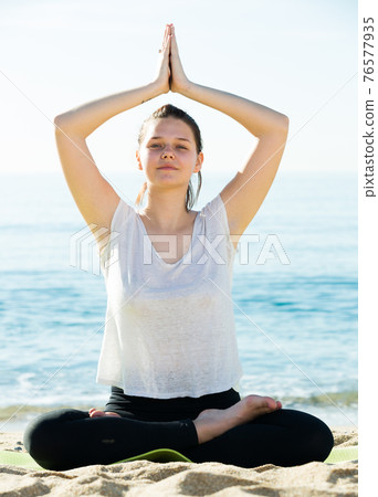 Smiling adult female in white T-shirt is sitting and practicing hatha Smiling adult female in white T-shirt is sitting and practicing hatha 76577935