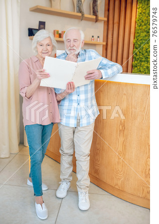 Gray-haired couple standing near reception in a spa center and looking trhough the price list Gray-haired couple standing near reception in a spa center and looking trhough the price list 76579748