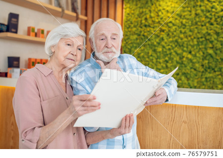 Gray-haired couple standing near reception in a spa center and looking trhough the price list Gray-haired couple standing near reception in a spa center and looking trhough the price list 76579751