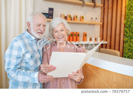 Gray-haired couple standing near reception in a spa center and looking trhough the price list Gray-haired couple standing near reception in a spa center and looking trhough the price list 76579757