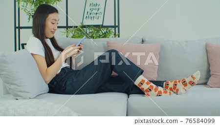 Young Asian girl chatting at social media networks use smartphone sitting on sofa. Mixed race woman relaxing on comfortable couch at home holding mobile. 76584099
