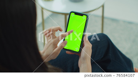 Back view of woman at smartphone with green screen for copy space. Chromakey mock up with tracking markers. Asian lady watching video news on couch close up. Tap to click on centre of screen. 76584107