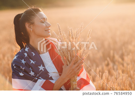 Woman with American flag and with a sheaf of ears in wheat field at sunset. 4th of July. Independence Day patriotic holiday. 76584950