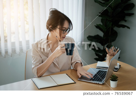 Portrait of beautiful accountant sitting at desk with interior drinking hot beverage holding cup with coffee looking at laptop while video conference. Portrait of beautiful accountant sitting at desk with interior drinking hot beverage holding cup with coffee looking at laptop while video conference. 76589067