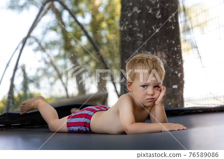 Portrait of cute bored little caucasian funny sad blond toddler boy lying inside big black trampoline at home backyard playground area outdoors warm summer sunny day. Children street sport activity 76589086