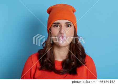 Young lady blowing bubble gum isolated over blue background, happy girl with dark hair with burst bubble of gum in mouth, wearing orange jumper and cap, looking at camera, looks funny. 76589432