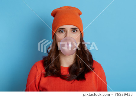 Young pretty lady with big eyes holding big chewing gum inside mouth, blowing large bubble, wearing cap and sweater, posing isolated over blue background. 76589433