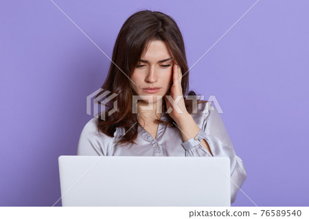 Young girl sitting in front of white laptop with exhausted facial expression, looks at pc screen, touching her face, having troubles with work, wearing white blouse, poses against lilac wall. Young girl sitting in front of white laptop with exhausted facial expression, looks at pc screen, touching her face, having troubles with work, wearing white blouse, poses against lilac wall. 76589540